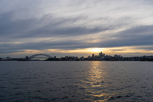 The Sydney skyline is silhouetted against a dramatic sunset, with golden hues reflecting on the harbor. The clouds add depth and contrast to the cityscape