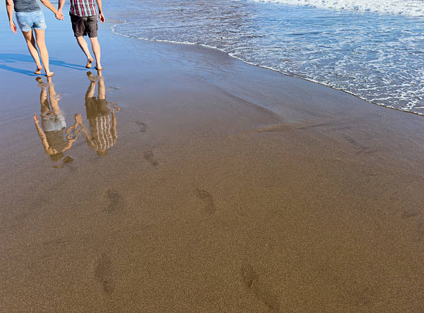 A close-up, low-angle shot of a couple walking barefoot and holding hands along the edge of the ocean on a wet, dark sand beach. The image captures their reflections clearly in the water-slicked sand. The foamy white edge of the surf rolls in alongside them, contrasting with the dark sand and bright sunlight, creating a feeling of romance, tranquility, summer vacation, and togetherness.