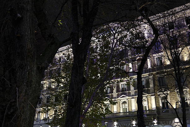 Night view of illuminated historic buildings and architectural details in an urban environment. Warm artificial lighting highlights classical facades, decorative elements, and street lamps against the dark sky, creating a dramatic and elegant atmosphere. Ideal for concepts of city life, travel, culture, heritage, nightlife, and European architecture