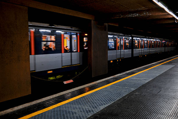 A modern subway train passes through an underground station with motion blur, while the platform and yellow safety line remain sharply in focus. Interior lights reveal passengers inside the moving train. The image conveys public transportation, urban mobility, commuting, and the fast-paced rhythm of city life at night.