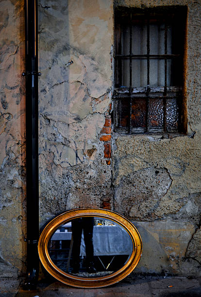 Old oval framed mirror resting on the ground against a cracked, weathered wall with a barred window. Reflections create a mysterious urban atmosphere, ideal for concepts of memory, reflection, decay, and abandoned architecture.