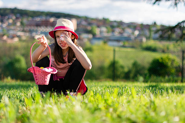 Young girl wearing a hat, crouching in grass, holding an Easter basket and an egg, celebrating spring