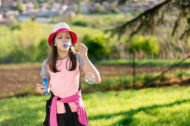 Girl in straw hat blowing soap bubbles in sunny backyard, joyful childhood play and carefree spring fun outdoors, vibrant sunlight