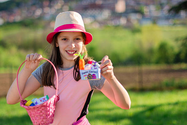 Young girl smiling and holding a pink Easter basket and a small gift box with happy Easter message at sunset