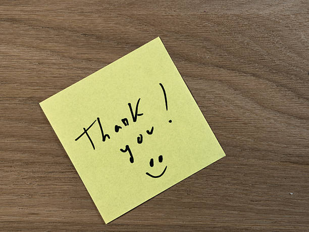 istock Handwritten Thank You on Yellow Sticky Note - Bright yellow sticky note with a handwritten 'Thank you!' message and smiley face on a warm, textured wooden desk background. Expressing gratitude. 2249300551