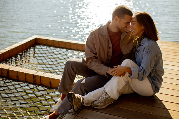 A couple shares an intimate moment by the water, exchanging smiles and laughter as the sun sets behind them, creating a warm glow.