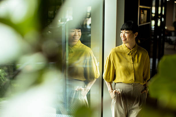 istock Woman in yellow blouse stands by large window in a modern workspace during daylight 2249303936