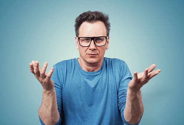 istock Confused man in glasses raising his hands in a questioning gesture, expressing frustration or disbelief. Casual blue t-shirt, puzzled facial expression. On blue background. 2249306665