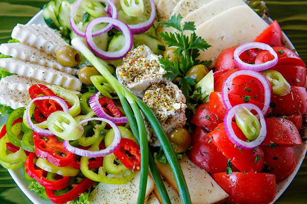 Sliced pepper, onion, tomato, cucumber, lettuce leaves, scallion, green olives and three types of cheese on appetizer plate on restaurant table, garnished with parsley and basil