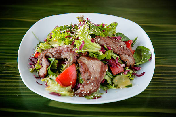 Grilled veal slices salad with romaine lettuce, spinach and tomato chops, sesame seed on top, served in plate on the table