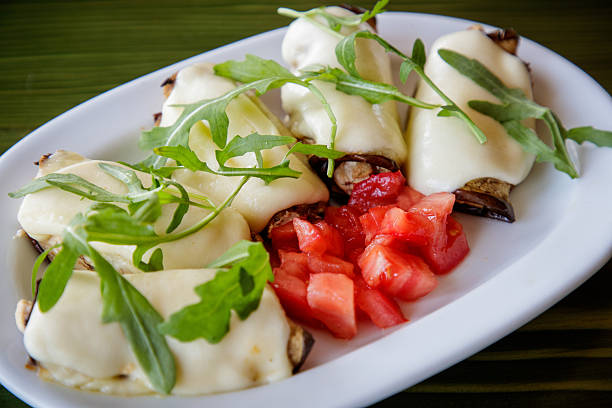 Chicken white meat rolled in eggplant slices and melted cheese, served with arugula and chopped tomato in plate on the table, close-up