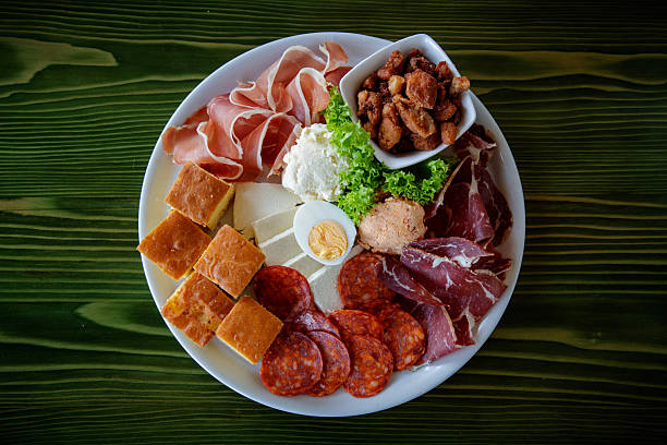 Directly above appetizer plate served on restaurant table, sliced ham, prosciutto, peperoni, boiled egg, corn bread, white and cottage cheese and pork rind