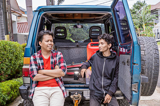 Two Indonesian southeast asian men relaxed and discussed while leaning on the open trunk of a blue car