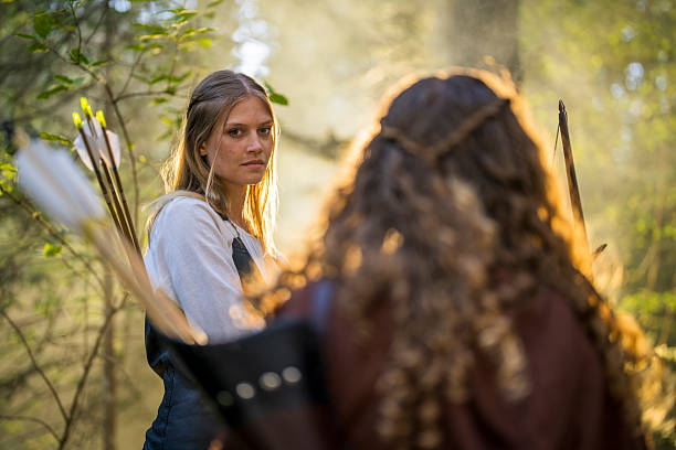 Young Caucasian female archers in a sunlit forest setting practicing archery with determined expressions. Surrounded by trees, they hold bows and arrows, concentrating on their medieval-themed activity.