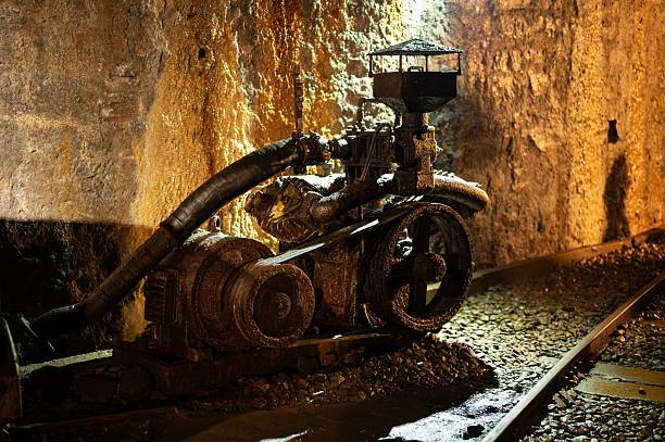 A heavily rusted, old twin-cylinder piston air compressor, driven by an electric motor, sits on rail tracks inside a dark coal mine tunnel.