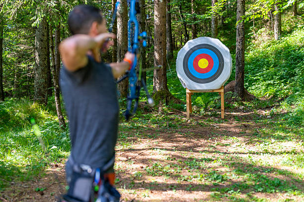 Mid adult Caucasian man in a forest concentrating on archery practice with a compound bow, aiming at a vibrant target while surrounded by lush trees and dappled sunlight.