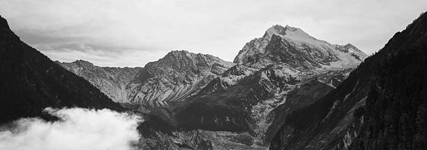 Epic monochrome landscape featuring a majestic, jagged snow-dusted summit dominating a deep glacial valley, framed by steep forested slopes and swirling low-lying clouds under a brooding sky. The high-contrast treatment accentuates the raw textures of rock faces, glacial ice, and dense coniferous forests, evoking timeless power, solitude, and the sublime beauty of untouched high-altitude wilderness. Ideal for themes of mountaineering, adventure travel, environmental serenity, meditation, or cinematic black-and-white photography. High-resolution, royalty-free stock image perfect for editorial covers, outdoor branding, large prints, book illustrations, or inspirational posters.