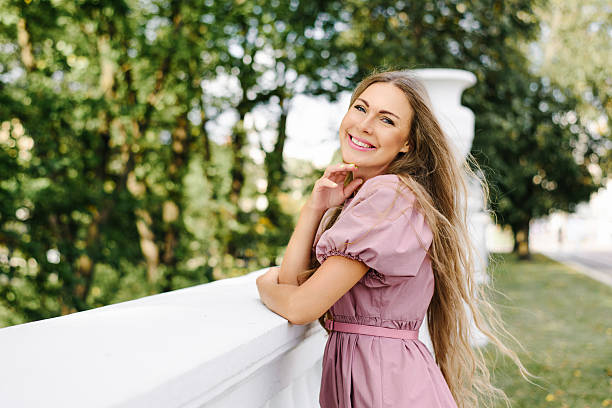 Happy young woman with long blonde hair and pink dress leaning on a white railing in a park, creating a cheerful and relaxed lifestyle scene.