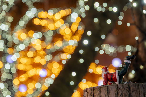 A small Santa Claus figurine is posed playfully on a tree stump, raising one leg as if stretching or exercising. Behind it, vibrant Christmas illumination creates large, colorful bokeh circles in warm yellow and cool white tones, producing a festive and whimsical holiday backdrop.