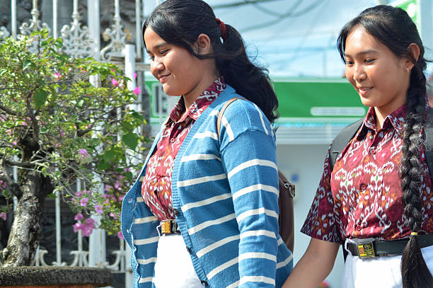 A close-up shows two cheerful students in patterned uniforms walking side by side holding hands near flowering plants, looking relaxed and happy as they arrive at the school compound.