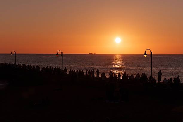 A silhouetted people gather along a waterfront at sunset, with a ship visible on the horizon under a vibrant orange sky.