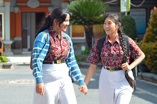 Two cheerful students in patterned uniforms with school bags walk side by side holding hands on the school grounds, smiling and chatting happily after classes have ended.