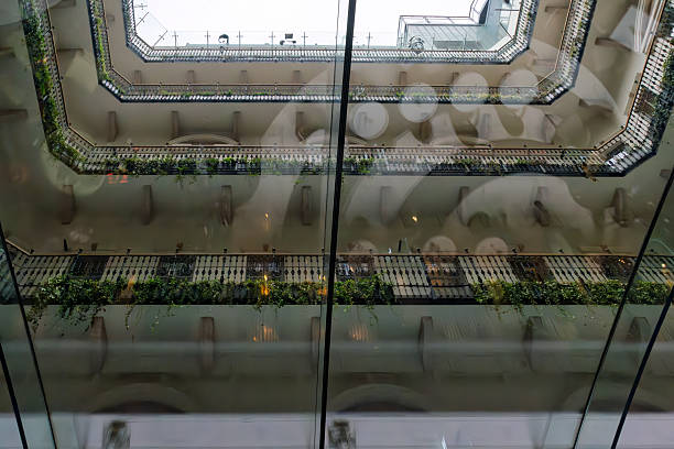 Budapest, HungaryA low-angle upward view captures a contemporary glass-enclosed atrium featuring a spiral staircase structure across multiple floors. Transparent glass walls and railings frame the vertical space, accented by lush green plants trailing along the edges and walkways.