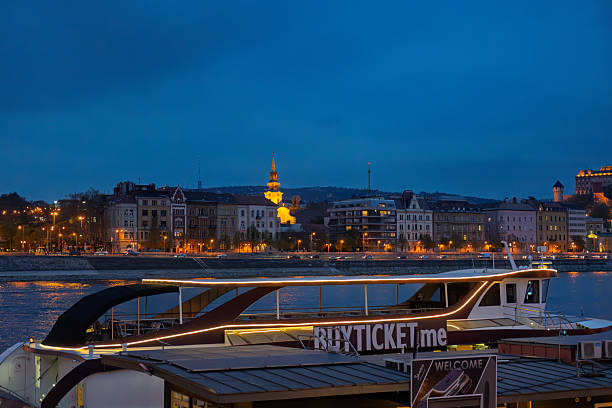 Budapest, Hungary, April  2025:The Buda Castle and Fisherman's bastion complex illuminated against the night sky, it has Baroque architecture featuring a prominent central dome and multiple towers highlighted in warm yellow and orange lights. In the foreground is a river cruise boat.