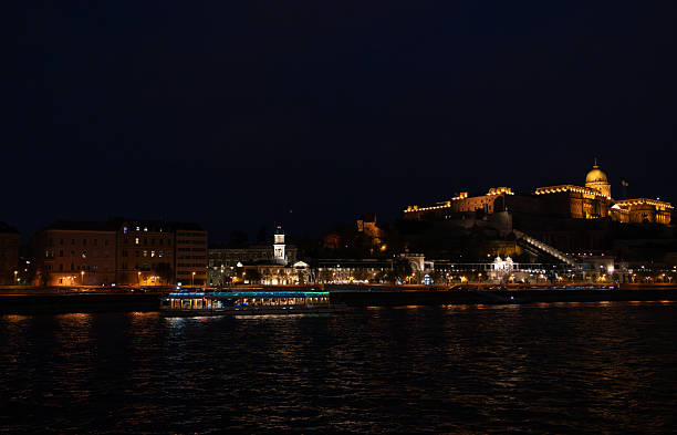 Budapest, Hungary, April  2025:The Buda Castle and Fisherman's bastion complex illuminated against the night sky, it has Baroque architecture featuring a prominent central dome and multiple towers highlighted in warm yellow and orange lights. The historic fortress, perched on the Buda hills, overlooks the Danube River, where reflections of the castle lights and surrounding street lamps can be seen on the surface. In the foreground, the river flows gently, bordered by riverside paths and trees. In the foreground is a river cruise boat.