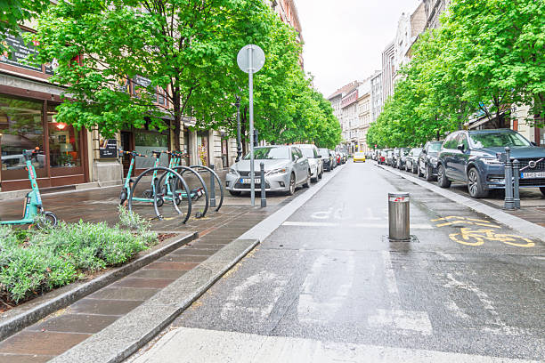 Budapest, Hungary, May 2025:A limited access road in the rain. There are electric scooters for hire on the left, a bollard in the road, cycle track signs and parked cars in this tree lined road.