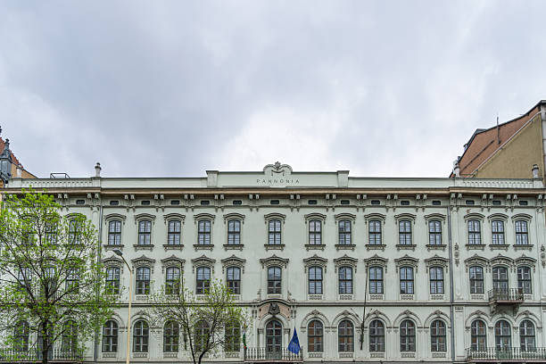Budapest, Hungary - May, 2025A grand historic building with intricate architectural details, including multiple arches, towers, and ornate facades, stands illuminated by warm lights against the overcast sky. The multi-story structure features large windows and decorative elements.  It has Pannonia on the roof top.