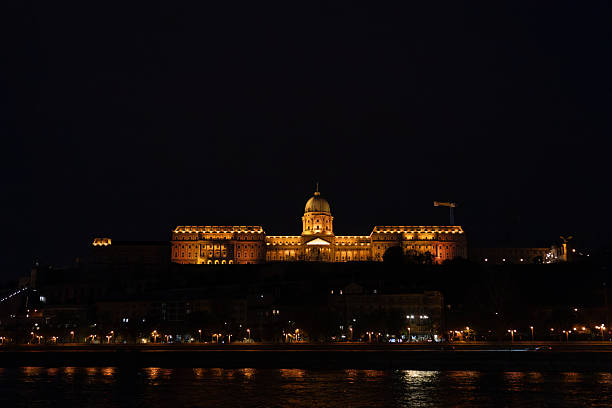Budapest, Hungary, April  2025:The Buda Castle and Fisherman's bastion complex illuminated against the night sky, it has Baroque architecture featuring a prominent central dome and multiple towers highlighted in warm yellow and orange lights. The historic fortress, perched on the Buda hills, overlooks the Danube River, where reflections of the castle lights and surrounding street lamps can be seen on the surface. In the foreground, the river flows gently, bordered by riverside paths and trees.