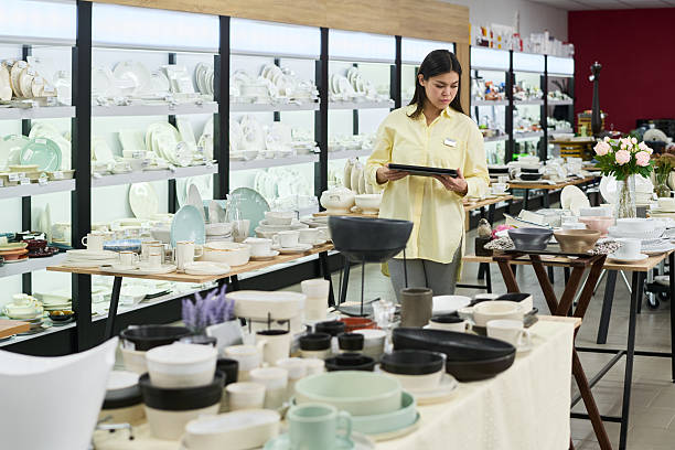 Young adult woman standing in tableware store holding digital tablet, checking inventory among shelves filled with plates, bowls, cups and various ceramic kitchenware items