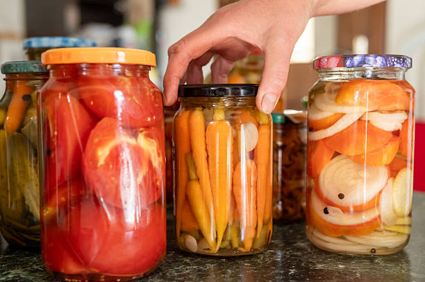 Hand reaching for pickled jars with assorted vegetables on kitchen counter.