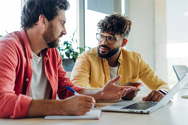 istock Smiling two businessmen sitting on workplace using laptop in modern office 2249347450