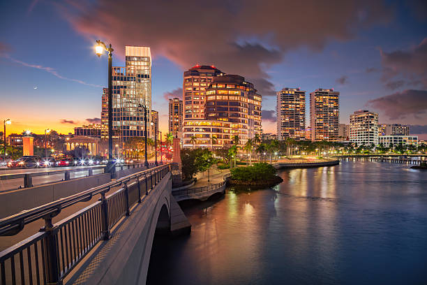 West Palm Beach, Florida, USA, during a sunset. The scene features a bridge leading into the city, bordered by a calm body of water. Modern high-rise buildings with illuminated windows stand prominently against a sky painted with warm hues of orange and purple. Streetlights line the bridge, casting reflections on the water below, while palm trees are visible along the waterfront, enhancing the tropical ambiance.