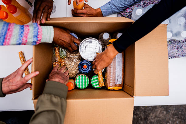 Close-up view of a collaborative community service moment, showing diverse hands working together to carefully place different food items and packaged goods into a cardboard box, symbolizing community support and collective care.