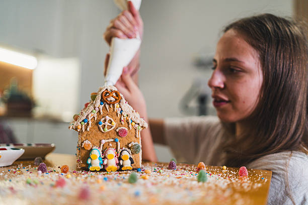 Woman carefully finishes a gingerbread house with colorful jelly candies and icing swirls. Cozy winter festive mood full of warmth, creativity and Christmas spirit.