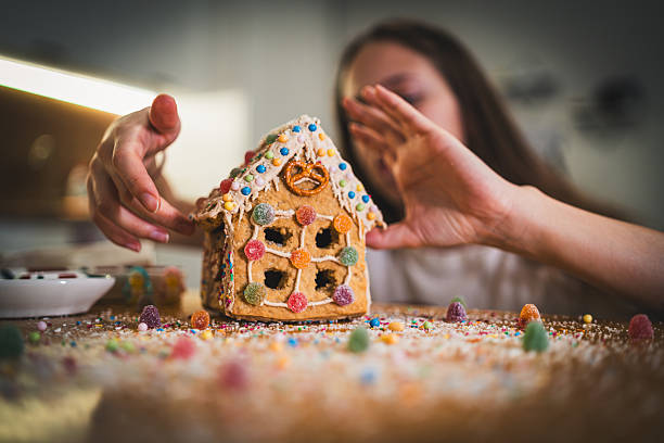 Festive moment of a woman designing a gingerbread house using icing and small candies. Soft lights, cozy winter atmosphere and holiday crafting tradition captured naturally.
