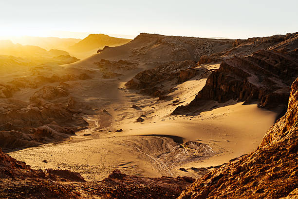 A dramatic desert landscape in the Atacama, Chile, bathed in warm golden light. Wind-sculpted dunes and rugged rock formations create a serene, sunlit scene perfect for travel and nature themes.