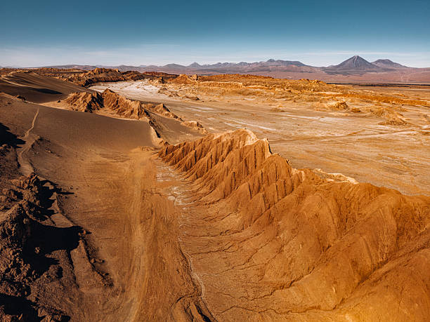 A wide, sunlit desert landscape in the Atacama of Chile features jagged dunes, rugged ridges, and distant volcanoes under a clear blue sky. Perfect for travel, nature, and geological themes.