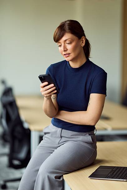 Focused Woman Using Smartphone At Modern Office Desk With Laptop In Neutral Space