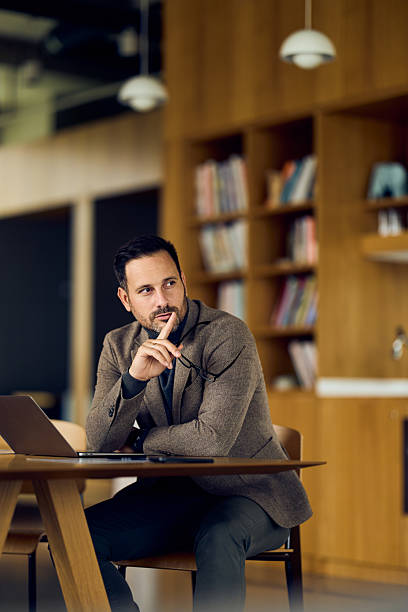 Thoughtful Professional Man In Brown Blazer At Conference Table In Modern Library Setting During Workday