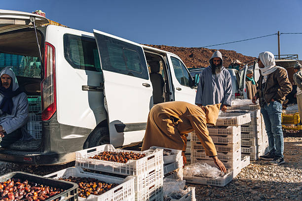 Erfoud, Morocco - February 15, 20125: Vibrant market activity in Erfoud, Morocco, with vendors handling crates of dates beside a white van while shoppers inspect goods under clear desert skies.