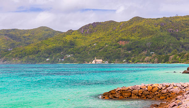 A serene tropical beach scene with turquoise water, a rocky breakwater, and lush green hills in the background. Anse Royale beach. Saint Joseph, the Worker church on the background. Seychelles