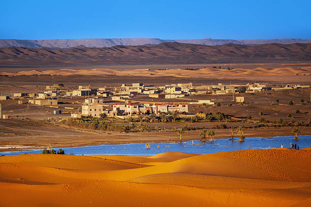 A desert village sits beside a bright blue oasis, with golden dunes in the foreground and distant mountains. Warm light highlights palm trees and adobe buildings, creating a tranquil Saharan landscape.