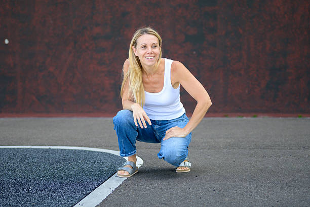 Happy blonde woman in casual white tank top and jeans squatting on pavement outdoors, relaxed and cheerful on a sunny day.