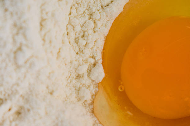 A top-down view of a raw egg placed in a well of flour. The flour is pale and finely milled, forming a circular mound around the egg. The egg yolk is a vibrant yellow-orange, surrounded by translucent egg white. This setup is commonly used in baking as a preparation step before mixing ingredients. The image focuses on the simplicity and purity of baking ingredients.