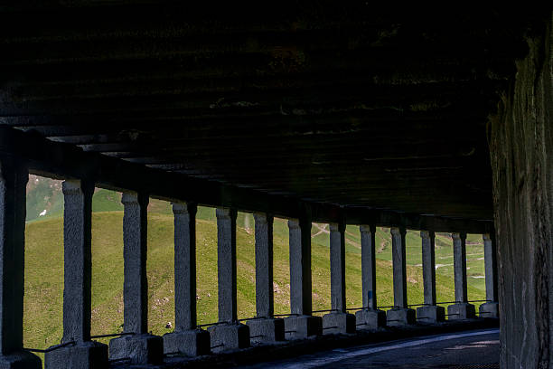 Dark covered road curves through mountain tunnel while sunlight grazes repetitive concrete support columns outside