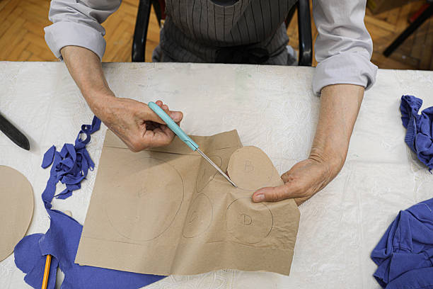 Close-Up View Of A Senior Woman (80+) Using Scissors To Cut Blue Fabric Along Paper Pattern Pieces On A Work Table In A Home Sewing Workshop, Illustrating Craftsmanship, DIY Creativity, And Active Aging.